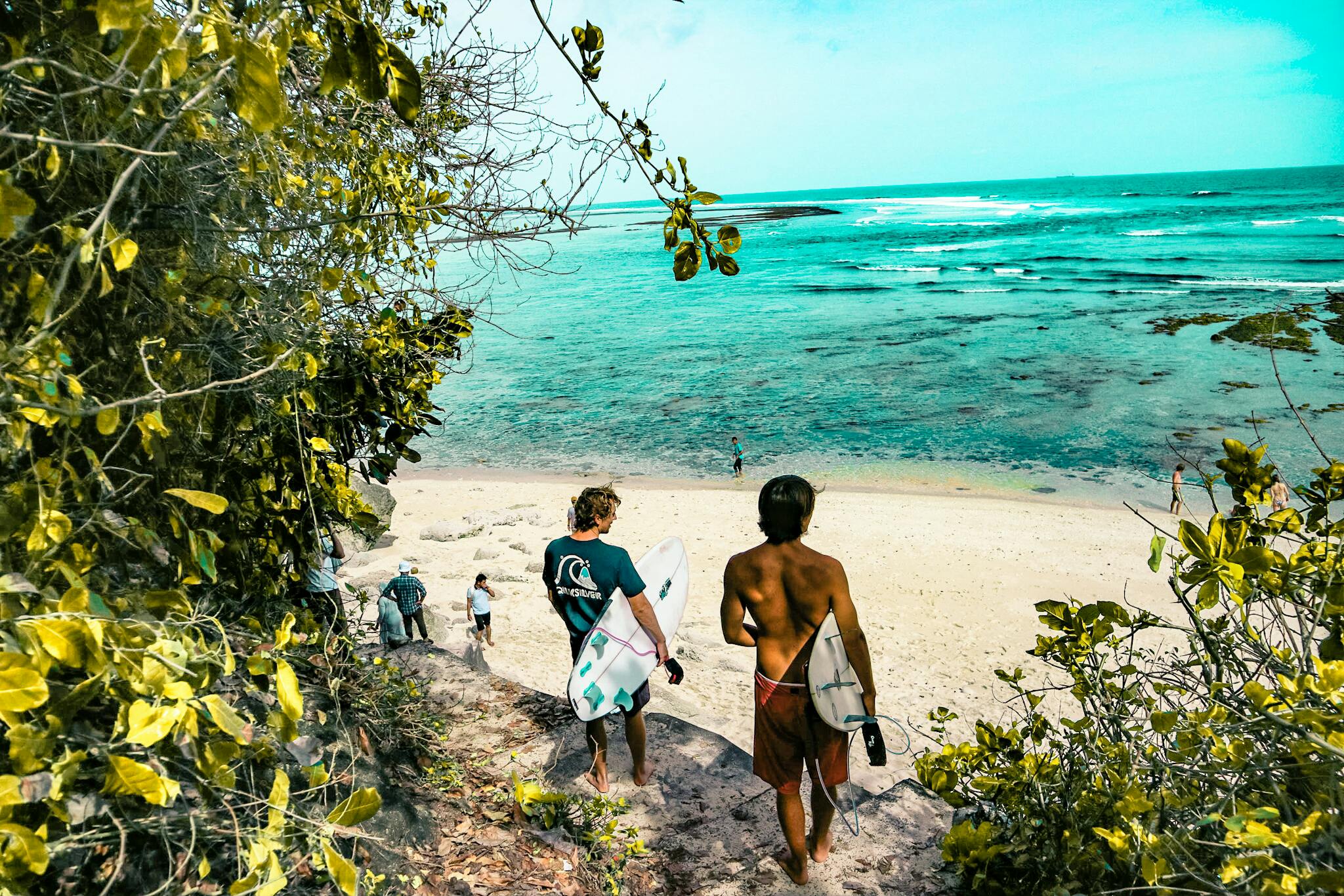 Surfers heading toward turquoise waves on a tropical Bali beach lined with palm trees